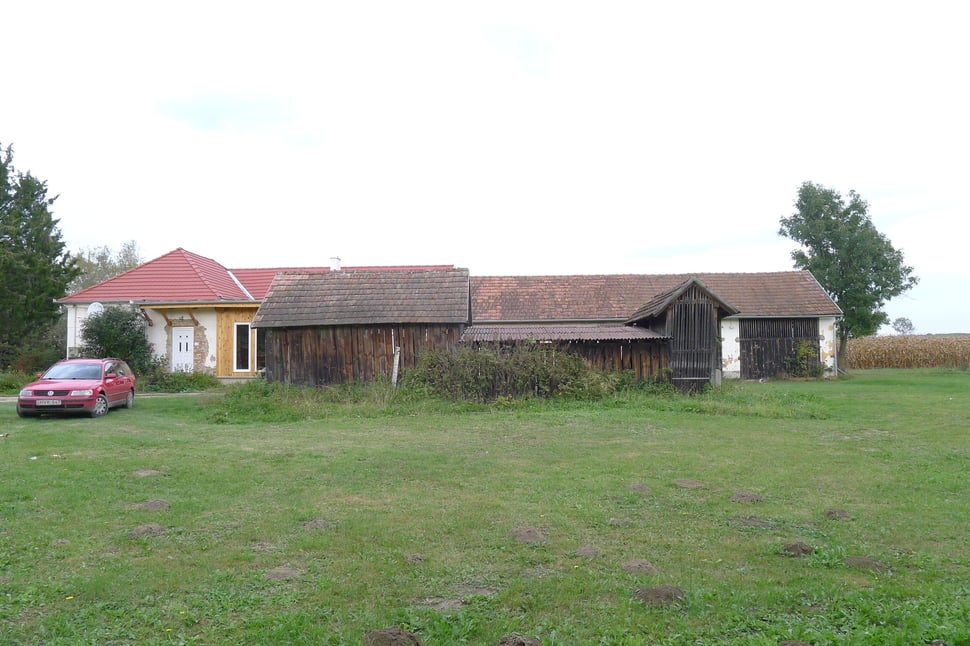 Old farm buildings and a lawn, with a red car parked at the left.