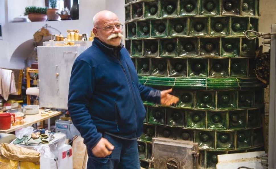 Péter Korompai in a blue jacket, standing beside stacks of green glass bottles in his workshop.