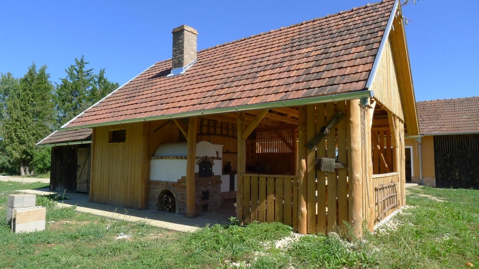 A wooden house with a tiled roof and open porch, seen across the grass.