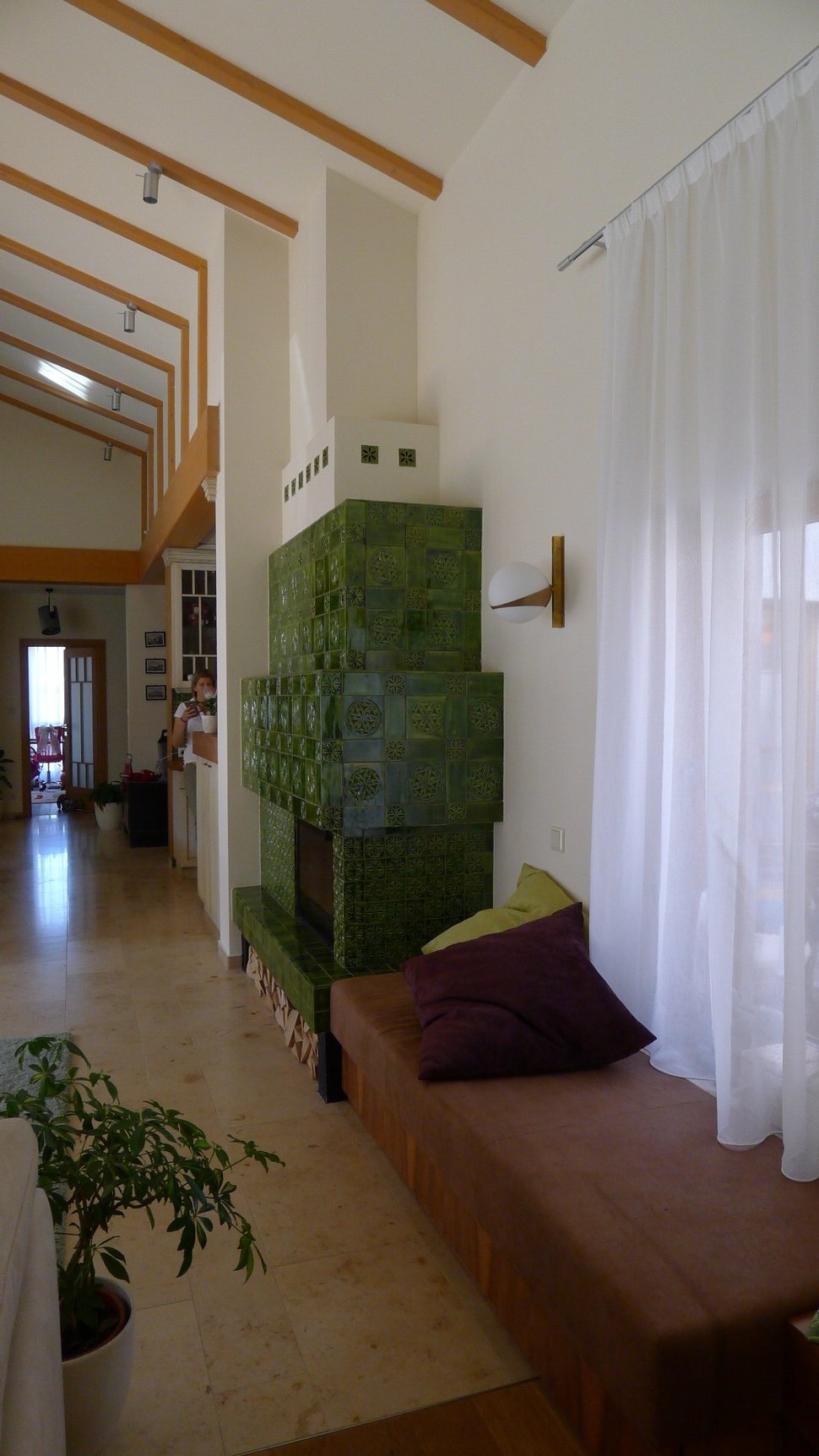 A green tiled stove in a narrow room with white curtains and a wooden bench.