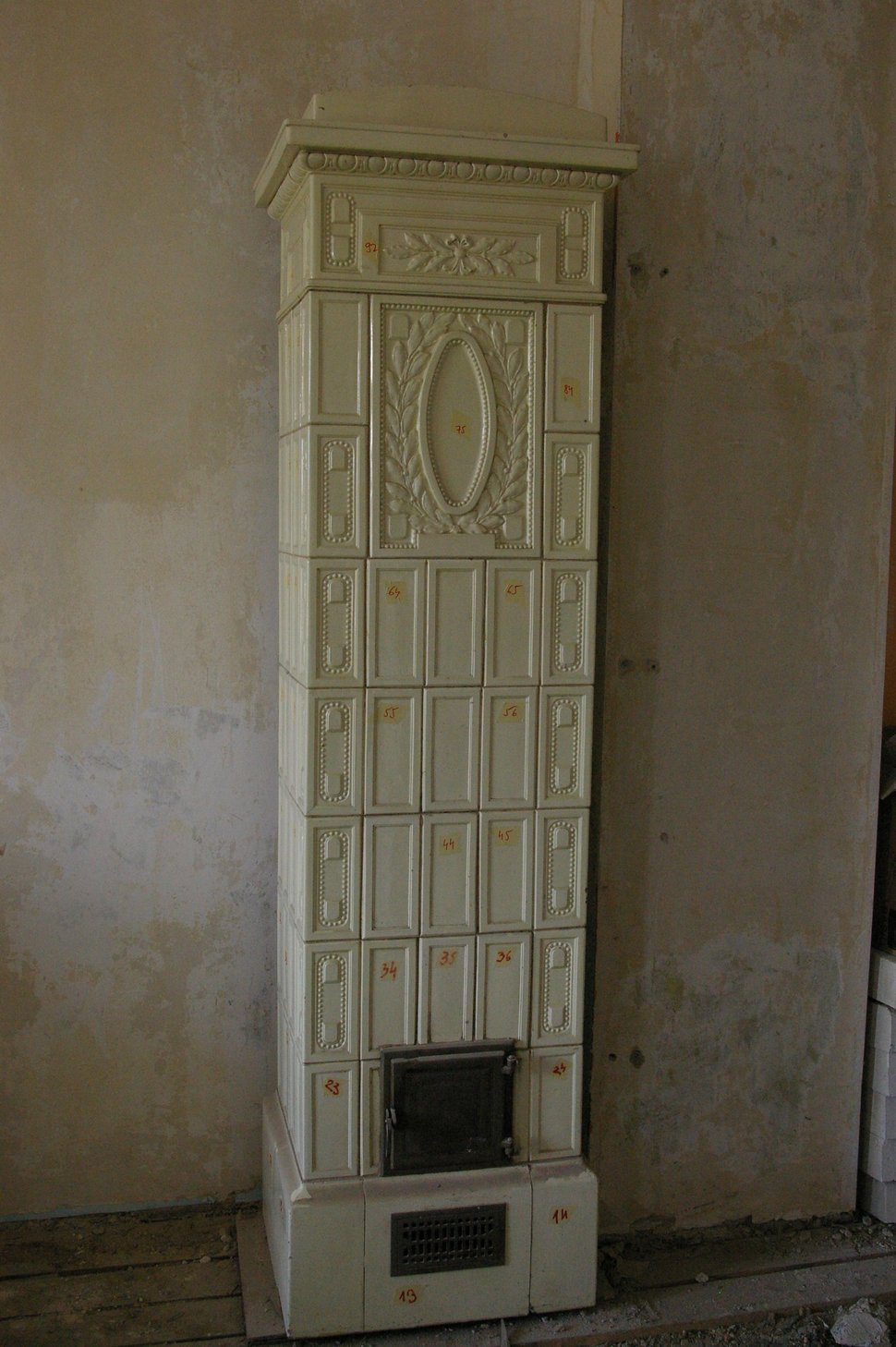 A tall cream tiled stove with relief panels standing against a bare wall.