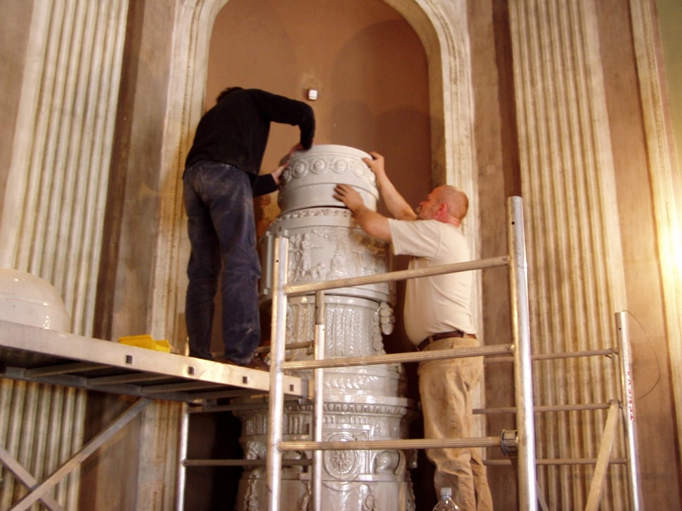 Two men assembling a tall white stove on scaffolding.