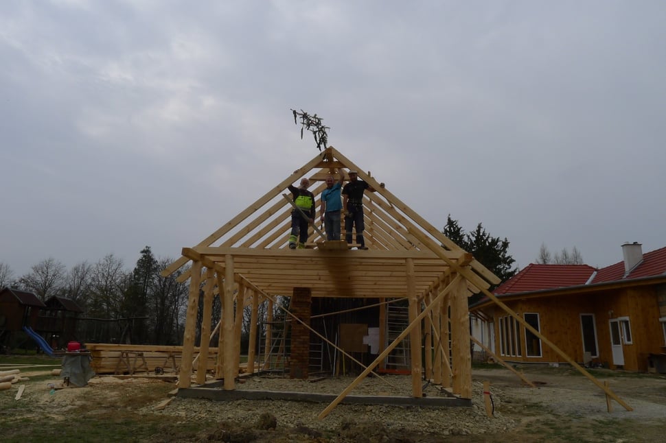 A timber frame roof being raised, with two men on top.