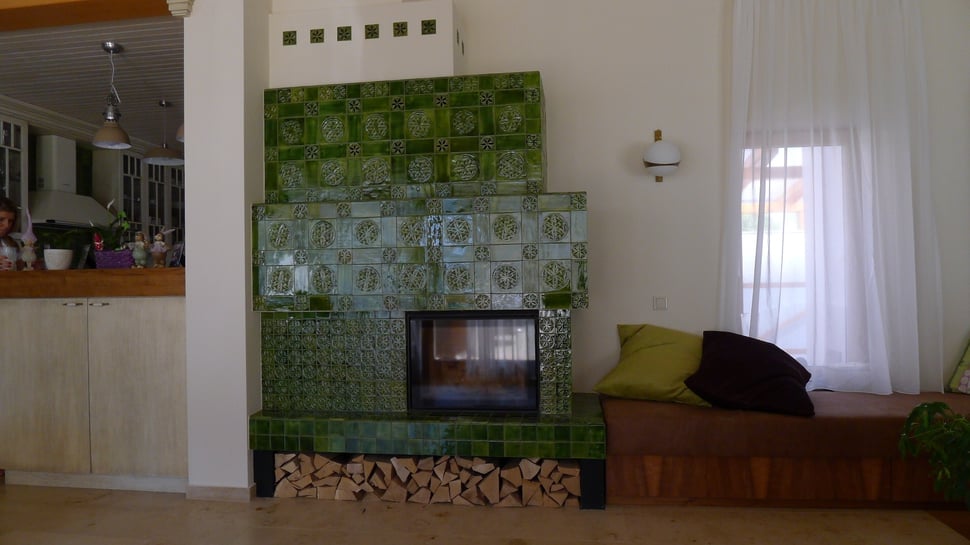 A green tiled stove in a white room, with firewood stored beneath it.