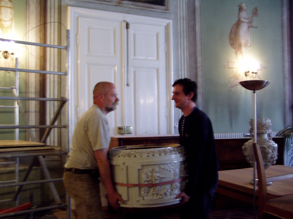 Two men carrying a round white stove section across a room.