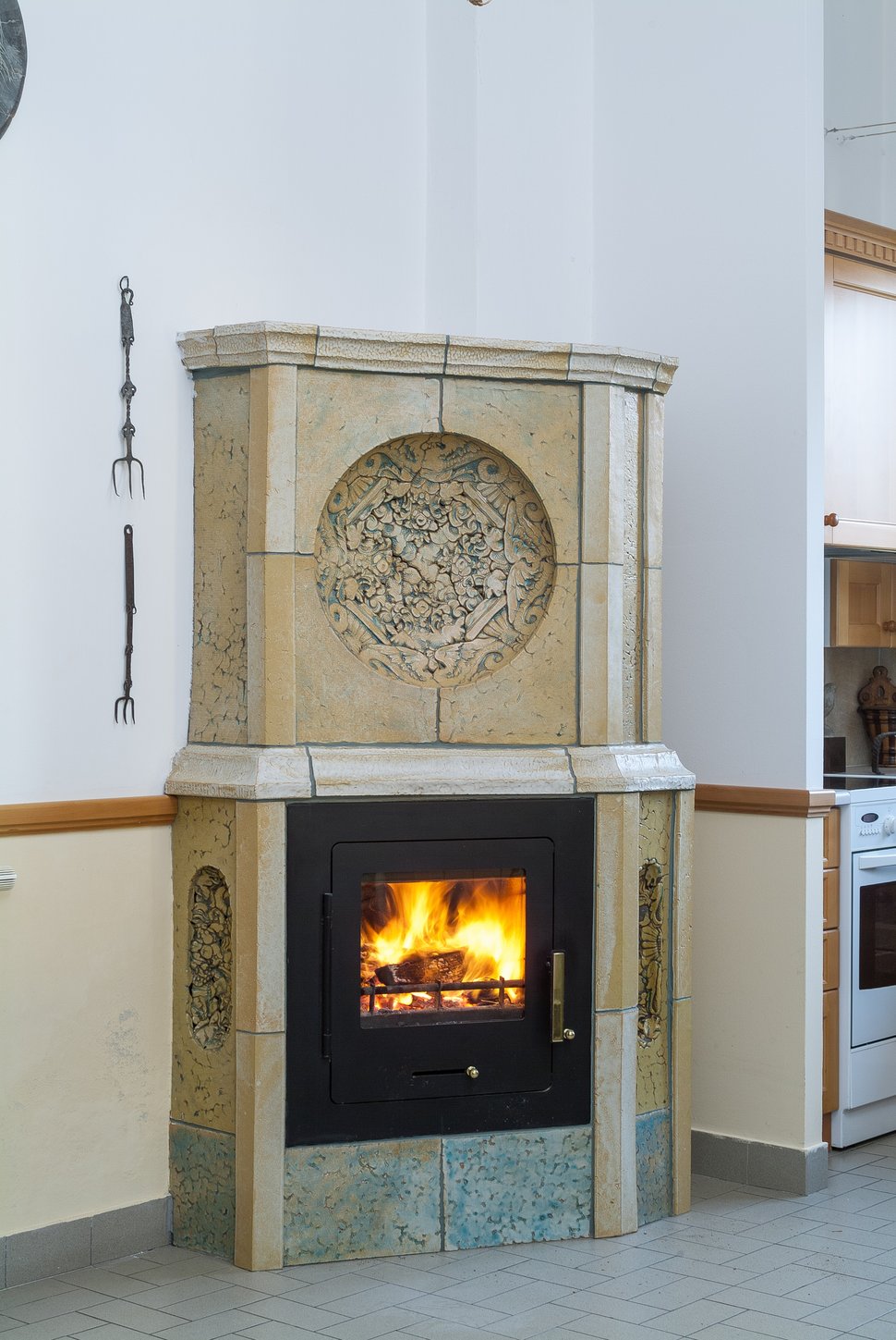 A tall pale tiled stove with a circular relief and black firebox in a white kitchen.