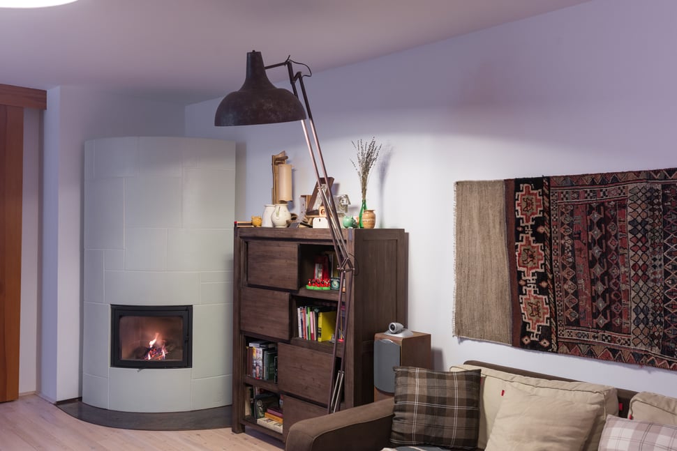 A pale tiled stove in a living room with a rug, bookshelf and lamp.